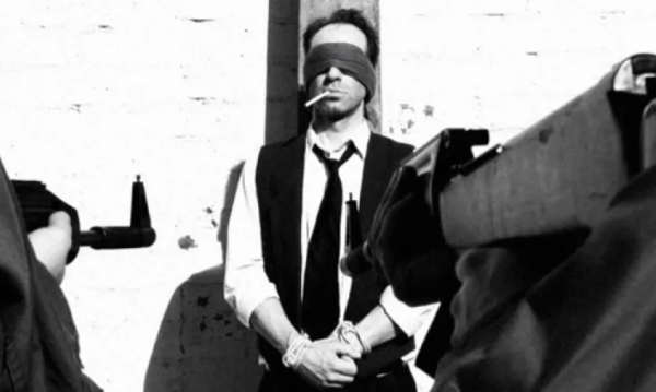 A man in blindfold, standing against a wall with a cigarette dangling from his mouth, as seen from the point of a firing squad.